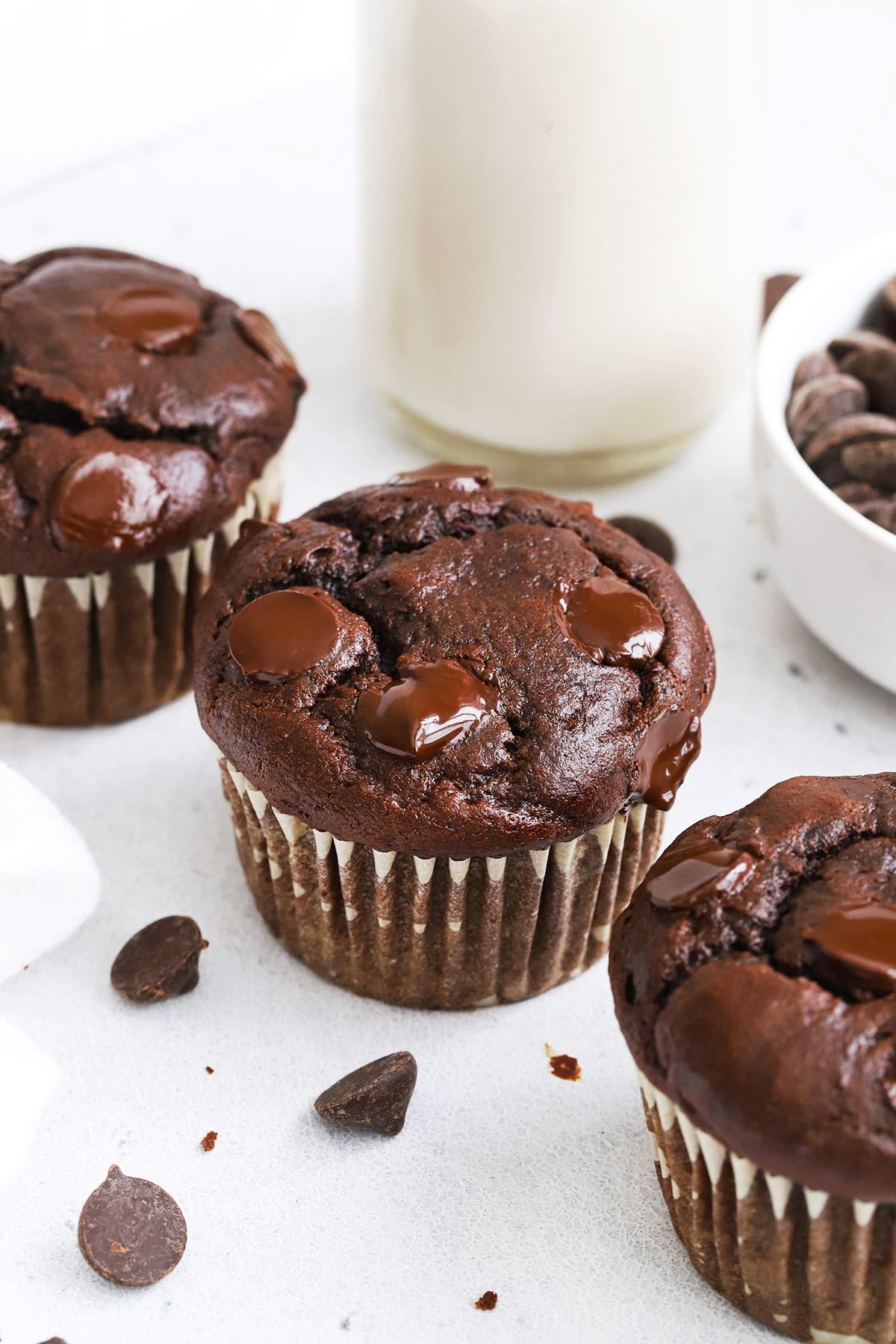 Three gluten-free chocolate banana muffins with a bowl of chocolate chips and a bottle of almond milk in the background