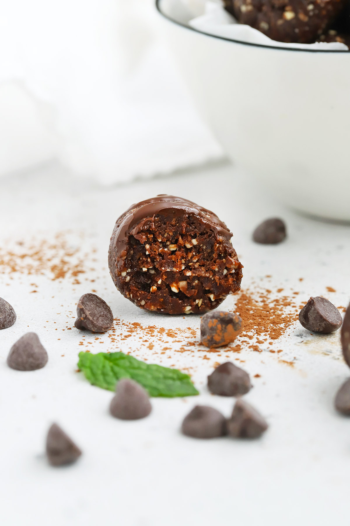 Close up front view of a mint chocolate energy bite with a bite taken out of it on a white background. Chocolate chips, cocoa powder, and mint leaves scattered around it. 