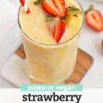Glass of Strawberry Orange Sunrise Smoothie topped with fresh pineapple and strawberries with a red and white stripe straw with text overlay that reads "paleo or vegan Strawberry Orange Sunrise Smoothie"