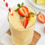Glass of Strawberry Orange Sunrise Smoothie topped with fresh pineapple and strawberries with a red and white stripe straw