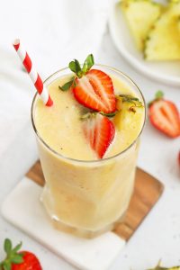 Glass of Strawberry Orange Sunrise Smoothie topped with fresh pineapple and strawberries with a red and white stripe straw