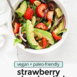 Overhead view of a white bowl of strawberry spinach salad and a fork on a white background with text overlay that reads "vegan + paleo-friendly strawberry spinach salad: fresh + light + bright"