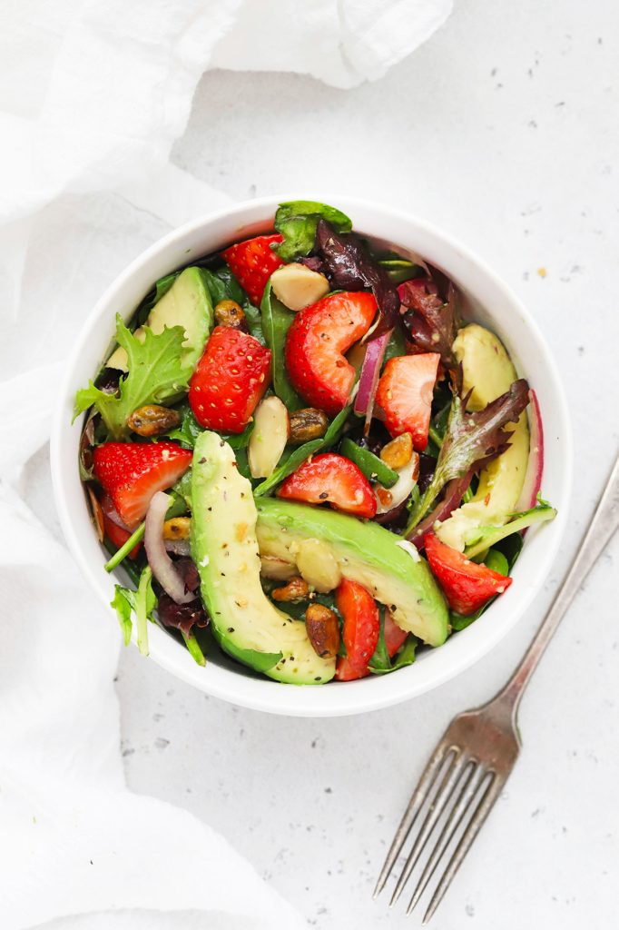Overhead view of a white bowl of strawberry spinach salad and a fork on a white background