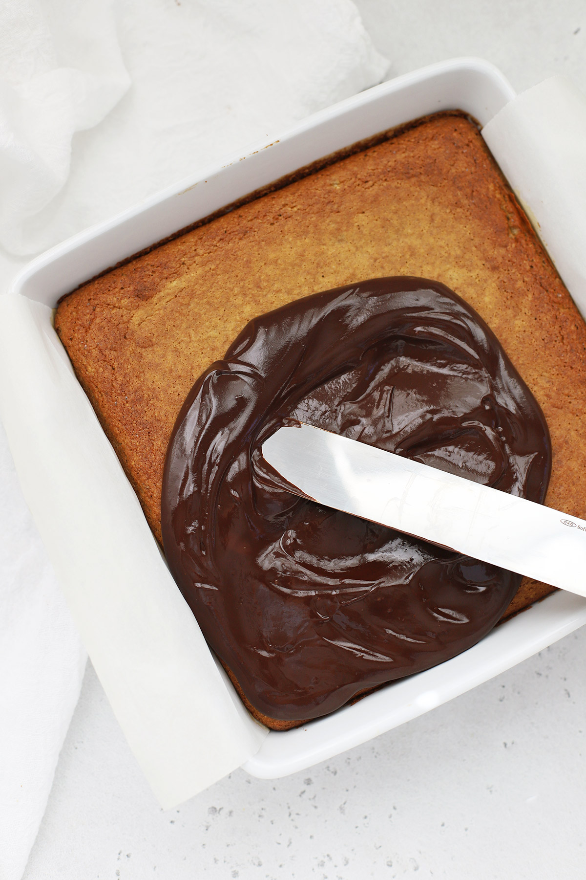overhead view of an offset spatula spreading chocolate ganache on an almond flour banana cake