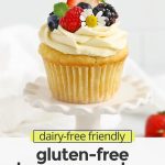 Front view of a gluten-free lemon cupcake with lemon frosting, fresh berries, and edible flowers on a white background with text overlay that reads "dairy-free friendly gluten-free lemon cupcakes: light + fluffy + irresistible!"