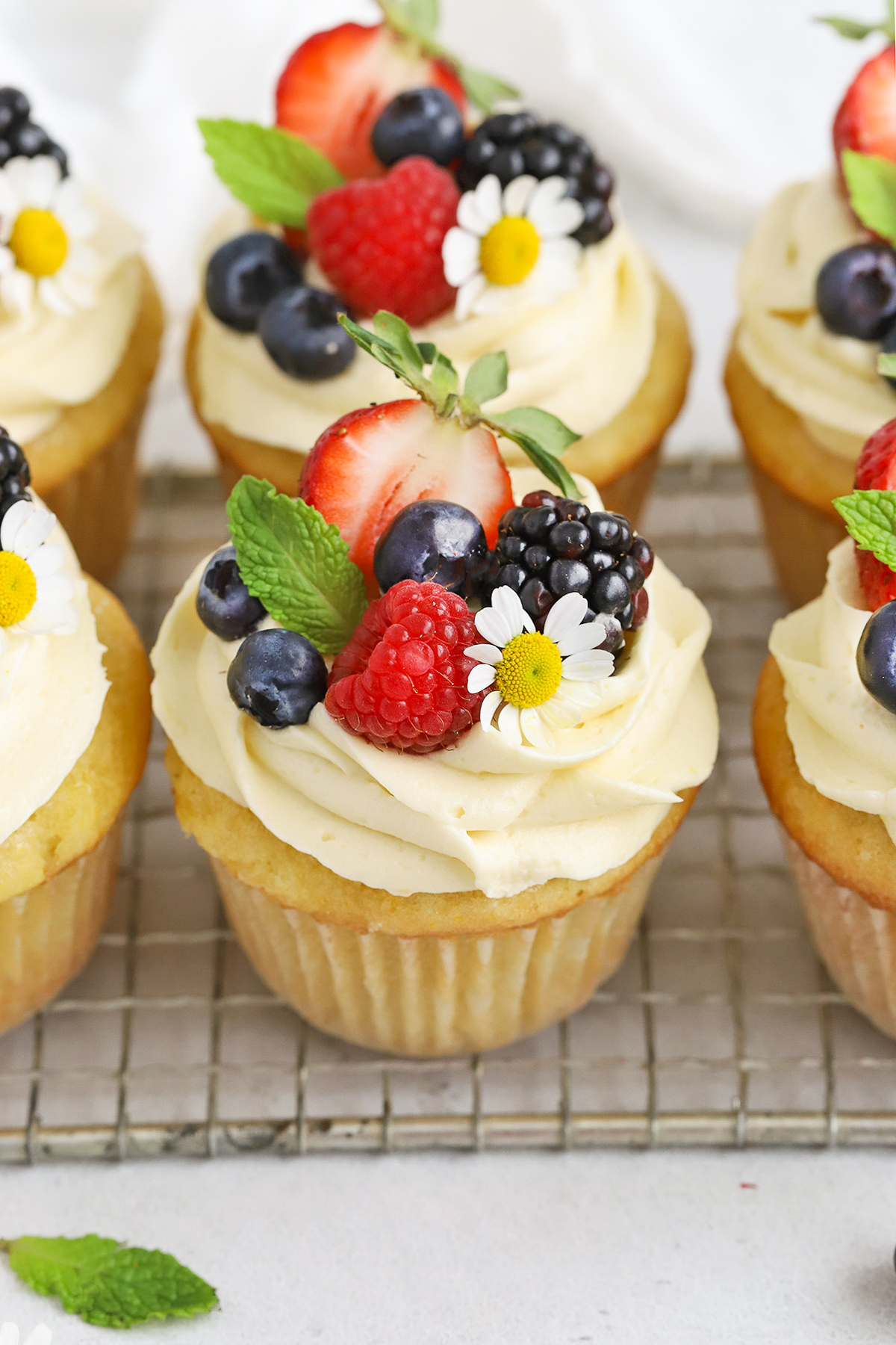 Front view of gluten-free lemon cupcakes with lemon frosting, fresh berries, and edible flowers on a white background