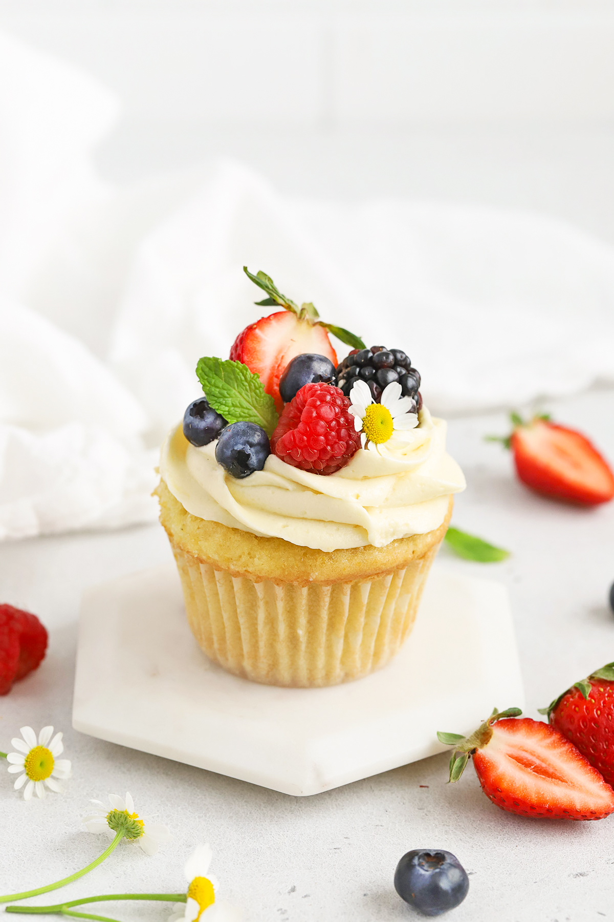 Front view of a gluten-free lemon cupcake with lemon frosting, fresh berries, and edible flowers on a white background