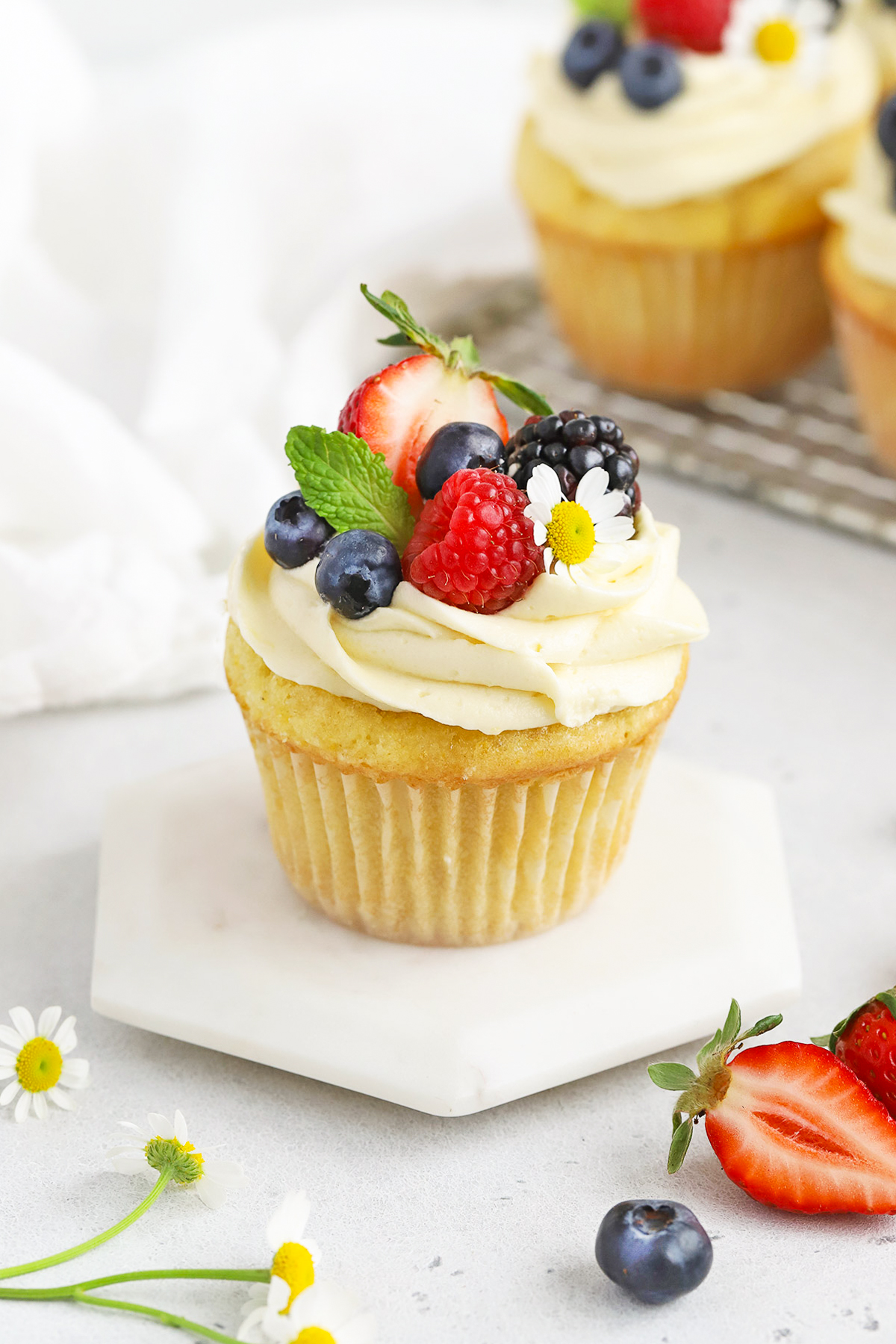 Front view of a gluten-free lemon cupcake with lemon frosting, fresh berries, and edible flowers on a white background