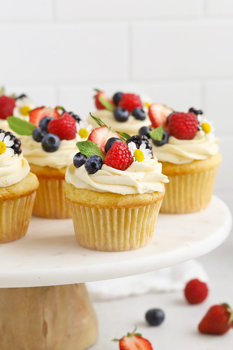 Front view of gluten-free lemon cupcakes with lemon frosting, fresh berries, and edible flowers on a white cake stand