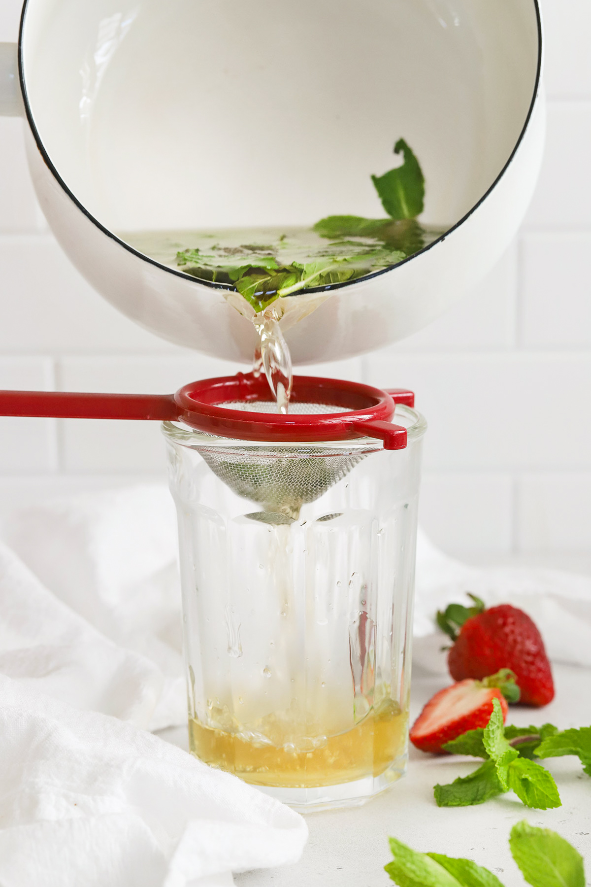 Front view of a saucepan of mint simple syrup being poured through a sieve into a glass container