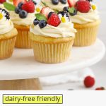 Front view of gluten-free lemon cupcakes with lemon frosting, fresh berries, and edible flowers on a white cake stand with text overlay that reads "dairy-free friendly gluten-free lemon cupcakes"