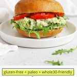 Front view of an avocado chicken salad sandwich with tomato and arugula on a white plate with text overlay that reads "gluten-free + paleo + whole30 Friendly Avocado Chicken Salad: Quick + Easy + Yummy"