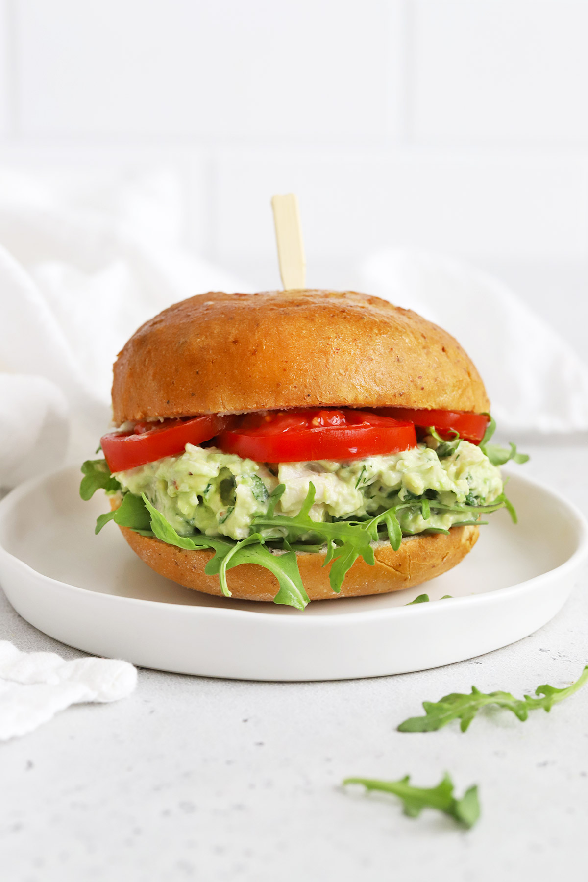 Front view of an avocado chicken salad sandwich with tomato and arugula on a white plate.