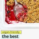 Overhead view of a baking dish of gluten-free berry crisp with a serving spoon with text overlay that reads "vegan-friendly. the best gluten-free berry crisp: gorgeous + easy + delicious"