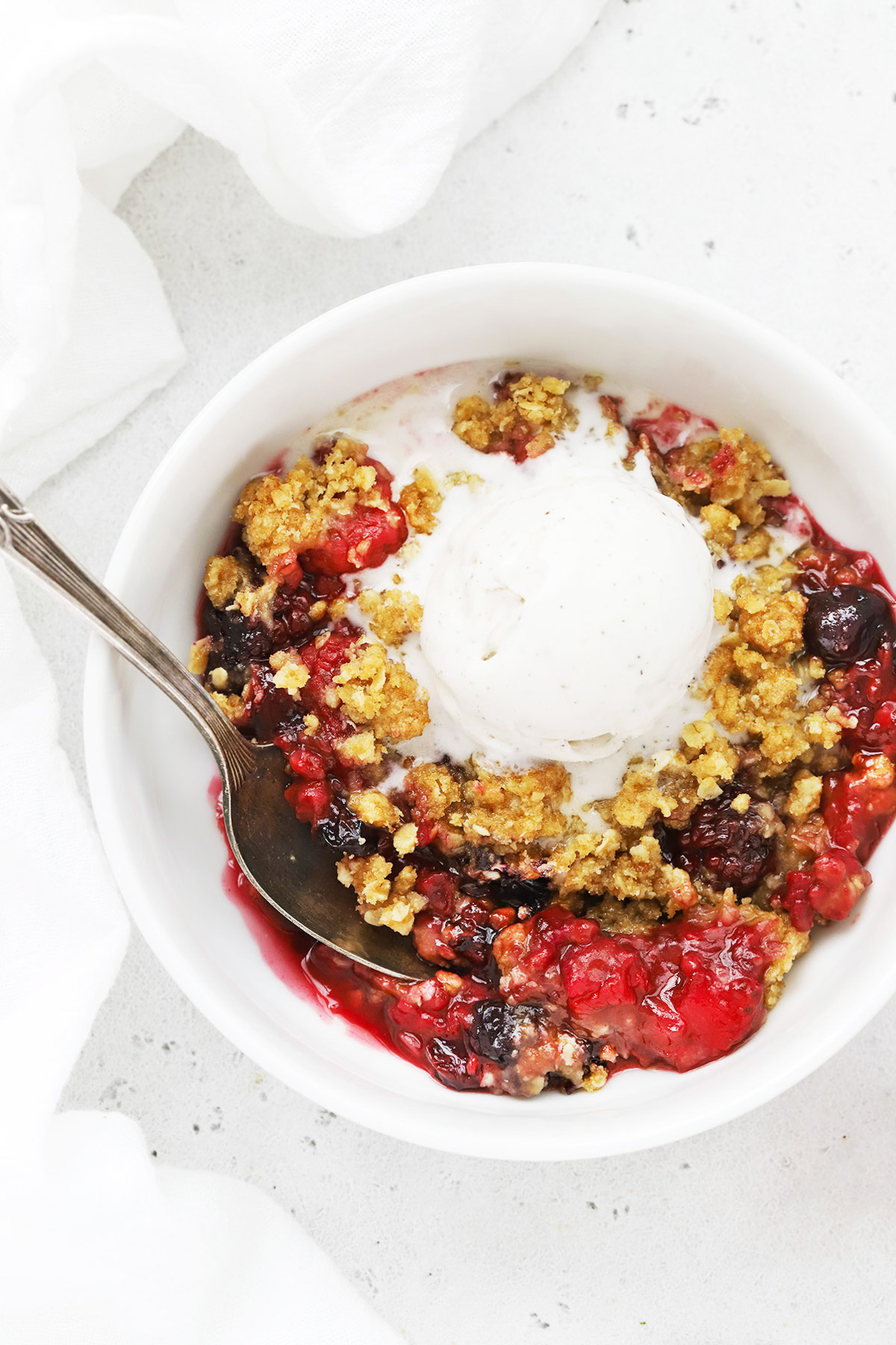 Close up overhead view of a bowl of gluten-free mixed berry crisp topped with dairy-free vanilla ice cream on a white background