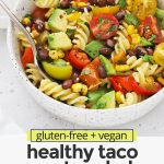Front view of a speckled bowl of healthy taco pasta salad on a white background with text overlay that reads "gluten-free + vegan healthy taco pasta salad with chili lime dressing"