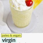 Overhead view of a virgin pina colada with whipped cream and a cherry on a white background with text overlay that reads "paleo & vegan virgin pina colada mocktails: quick + creamy + refreshing"