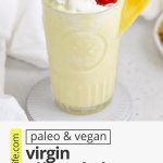 Front view of a virgin pina colada with whipped cream and a cherry on a white background with text overlay that reads "paleo & vegan virgin pina colada mocktails: quick + creamy + refreshing"