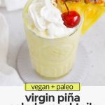 Overhead view of a virgin pina colada with whipped cream and a cherry on a white background with text overlay that reads "paleo & vegan virgin pina colada mocktails: quick + creamy + refreshing"