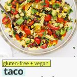 Overhead view of a glass mixing bowl of healthy taco pasta salad on a white background with text overlay that reads "gluten-free + vegan taco pasta salad with chili lime dressing: quick + healthy + fresh"
