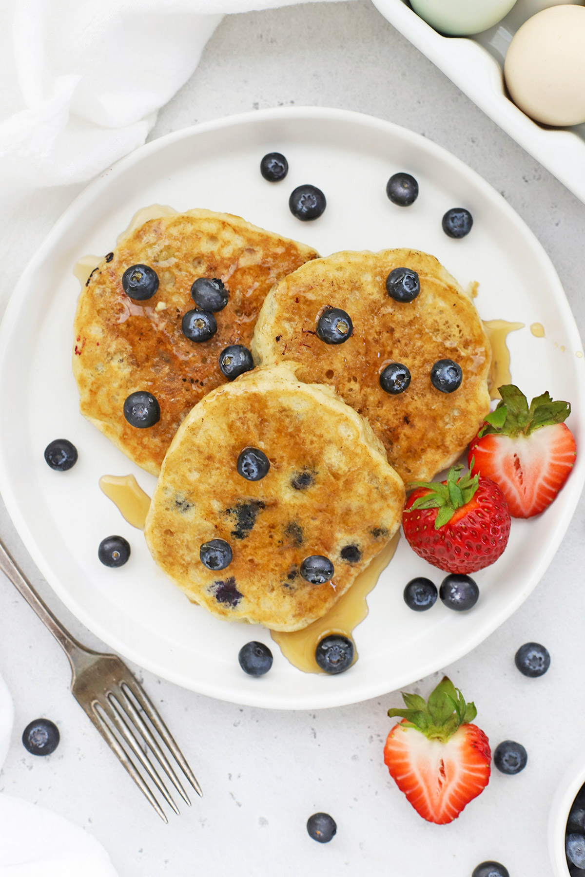 Overhead view of 3 fluffy gluten-free blueberry pancakes on a white plate drizzled with syrup
