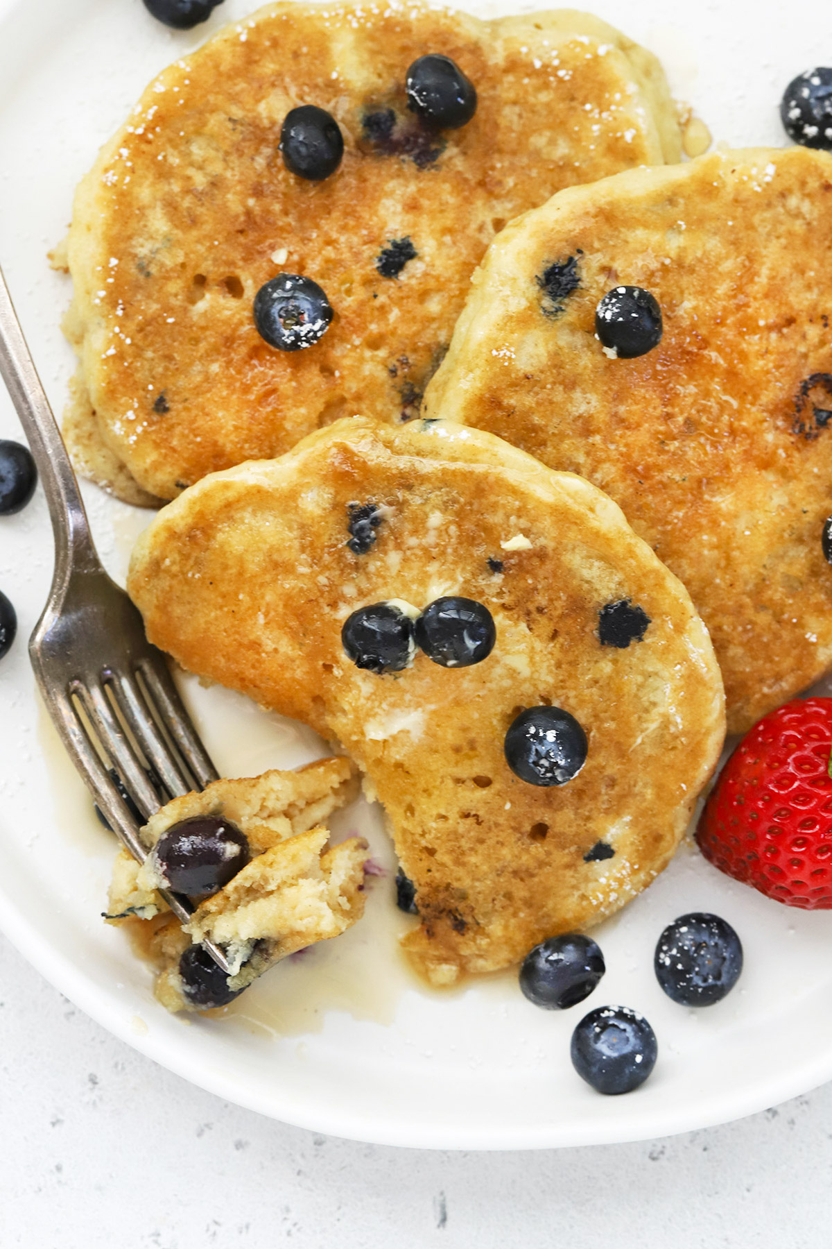 Close up view of 3 fluffy gluten-free blueberry pancakes on a white plate drizzled with syrup