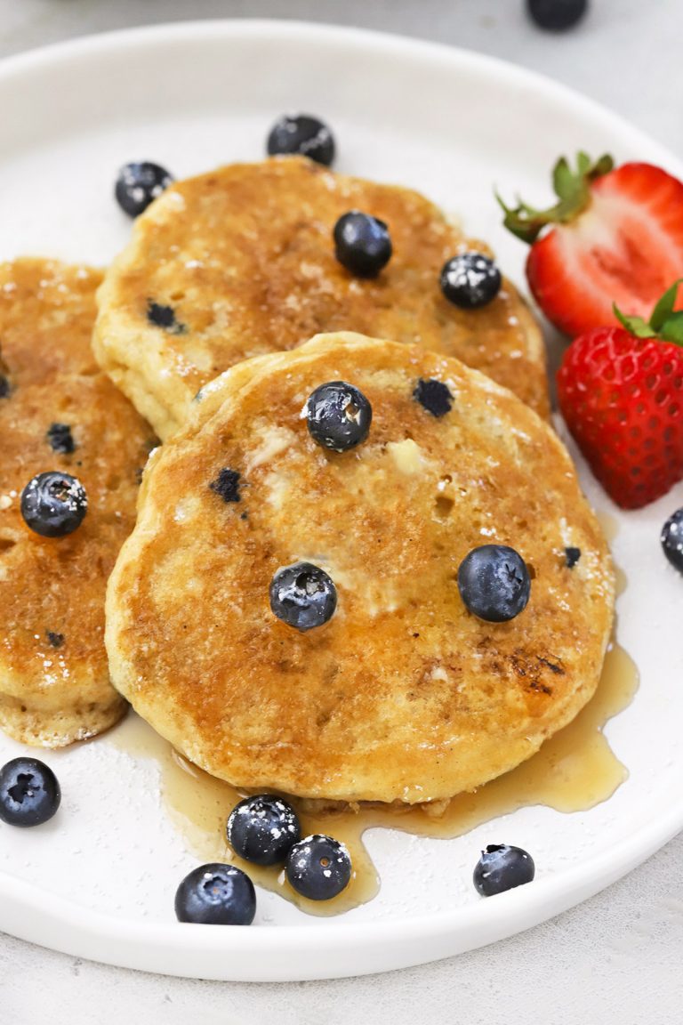 Front view of 3 fluffy gluten-free blueberry pancakes on a white plate drizzled with syrup