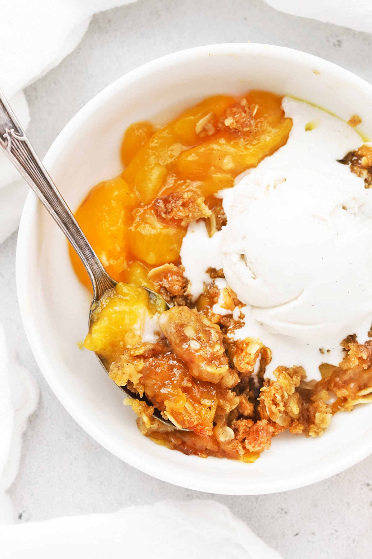 Close up overhead view of a bowl of gluten-free peach crisp topped with dairy-free vanilla ice cream, slowly melting over the warm crisp