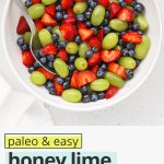 Overhead view of a white bowl of honey lime fruit salad with green grapes, strawberries, and blueberries with text overlay that reads "paleo + easy honey lime fruit salad: so fresh + simple + delicious!"