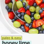 Close up overhead view of a white bowl of honey lime fruit salad with strawberries, blueberries, and green grapes with text overlay that reads "paleo + easy honey lime fruit salad: so fresh + simple + delicious!"