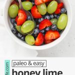 Close up overhead view of a small white bowl of honey lime fruit salad with strawberries, blueberries, and green grapes with text overlay that reads "paleo + easy honey lime fruit salad: so fresh + simple + delicious!"
