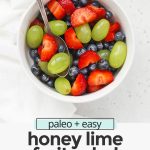 Close up overhead view of a small white bowl of honey lime fruit salad with strawberries, blueberries, and green grapes with text overlay that reads "paleo + easy honey lime fruit salad: so fresh + simple + delicious!"