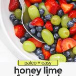 Close up overhead view of a white bowl of honey lime fruit salad with strawberries, blueberries, and green grapes with text overlay that reads "paleo + easy honey lime fruit salad: so fresh + simple + delicious!"