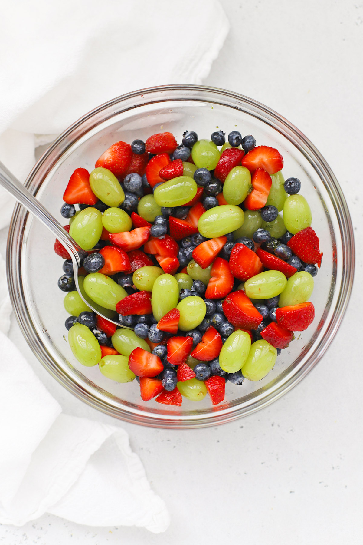 Front view of a white bowl of honey lime fruit salad with strawberries, blueberries, and green grapes freshly mixed in a glass bowl