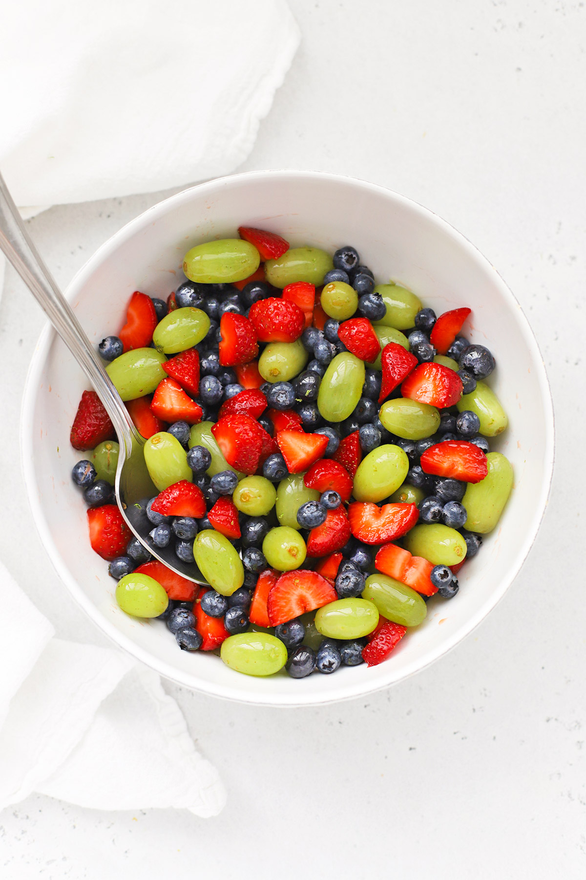 Overhead view of a white bowl of honey lime fruit salad with green grapes, strawberries, and blueberries.