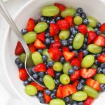 Close overhead view of a white bowl of honey lime fruit salad with green grapes, strawberries, and blueberries.