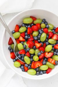 Close overhead view of a white bowl of honey lime fruit salad with green grapes, strawberries, and blueberries.