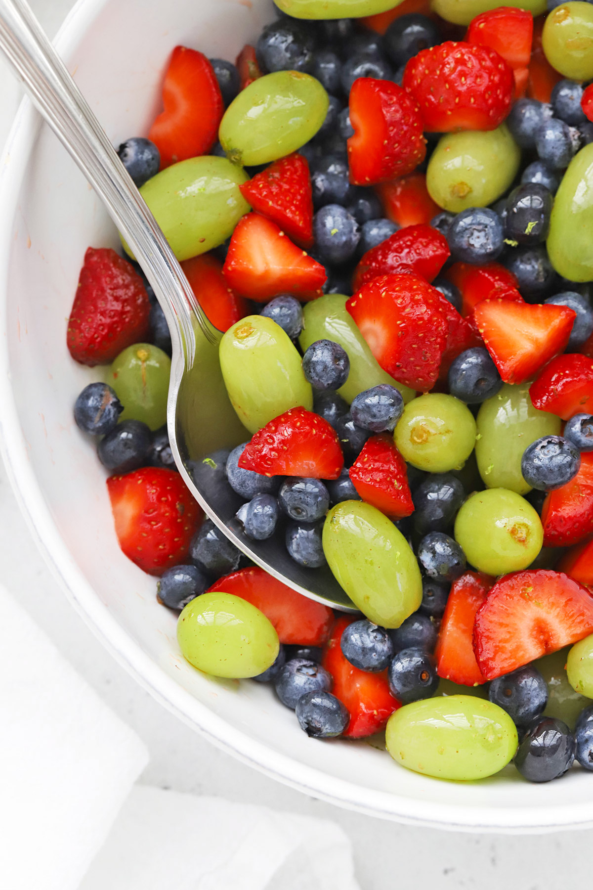 Close up overhead view of a white bowl of honey lime fruit salad with strawberries, blueberries, and green grapes.