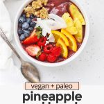 Overhead view of a pineapple acai bowl with fresh berries, pineapple, orange slices, shaved coconut, granola, mango on a white background with text overlay that reads "vegan + paleo pineapple acai bowls: fresh + beautiful + healthy"
