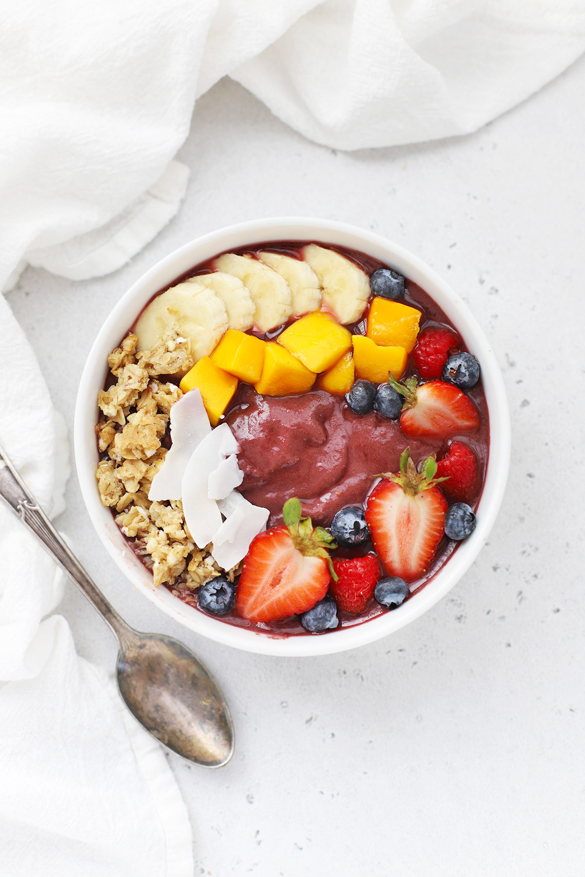 Overhead view of a pineapple acai bowl with fresh berries, shaved coconut, granola, mango, and banana on a white background