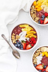 Overhead view of three pineapple acai bowls topped with fresh fruit and granola on a white background