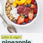 Overhead view of a pineapple acai bowl with fresh berries, shaved coconut, granola, mango, and banana on a white background with text overlay that reads "vegan + paleo pineapple acai bowls: fresh + beautiful + healthy"