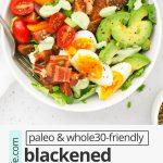 Overhead view of a blackened salmon cobb salad on a white background with avocado green goddess dressing on the side with text overlay that reads "paleo & whole30 blackened salmon cobb salad: A yummy healthy dinner"