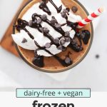 Overhead view of a glass of vegan frozen hot chocolate topped with coconut whipped cream and chocolate chips with text overlay that reads "dairy-free + vegan frozen hot chocolate: frosty + creamy + so delicious!"