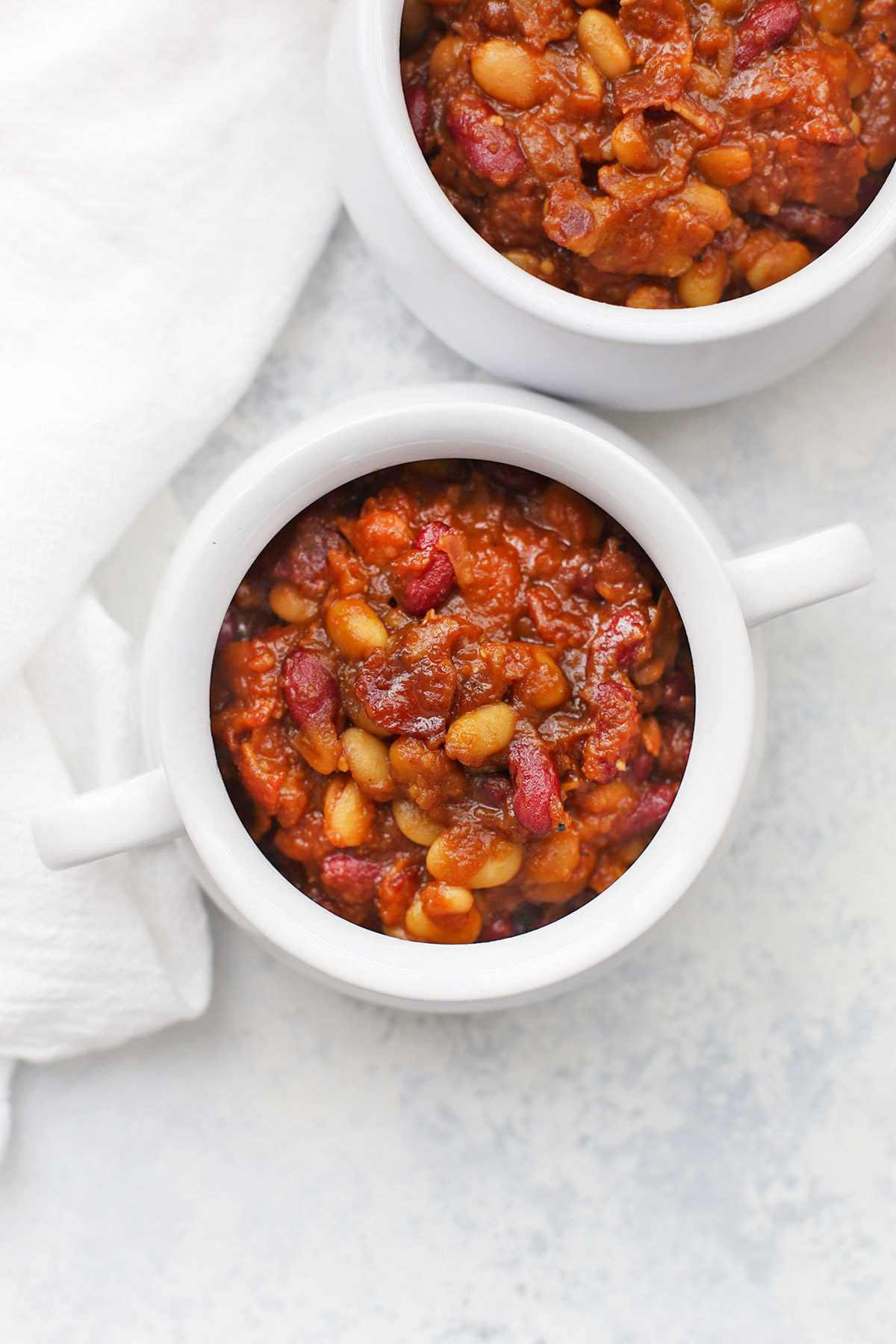 Overhead view of a white crock of settlers baked beans with bacon on a white background
