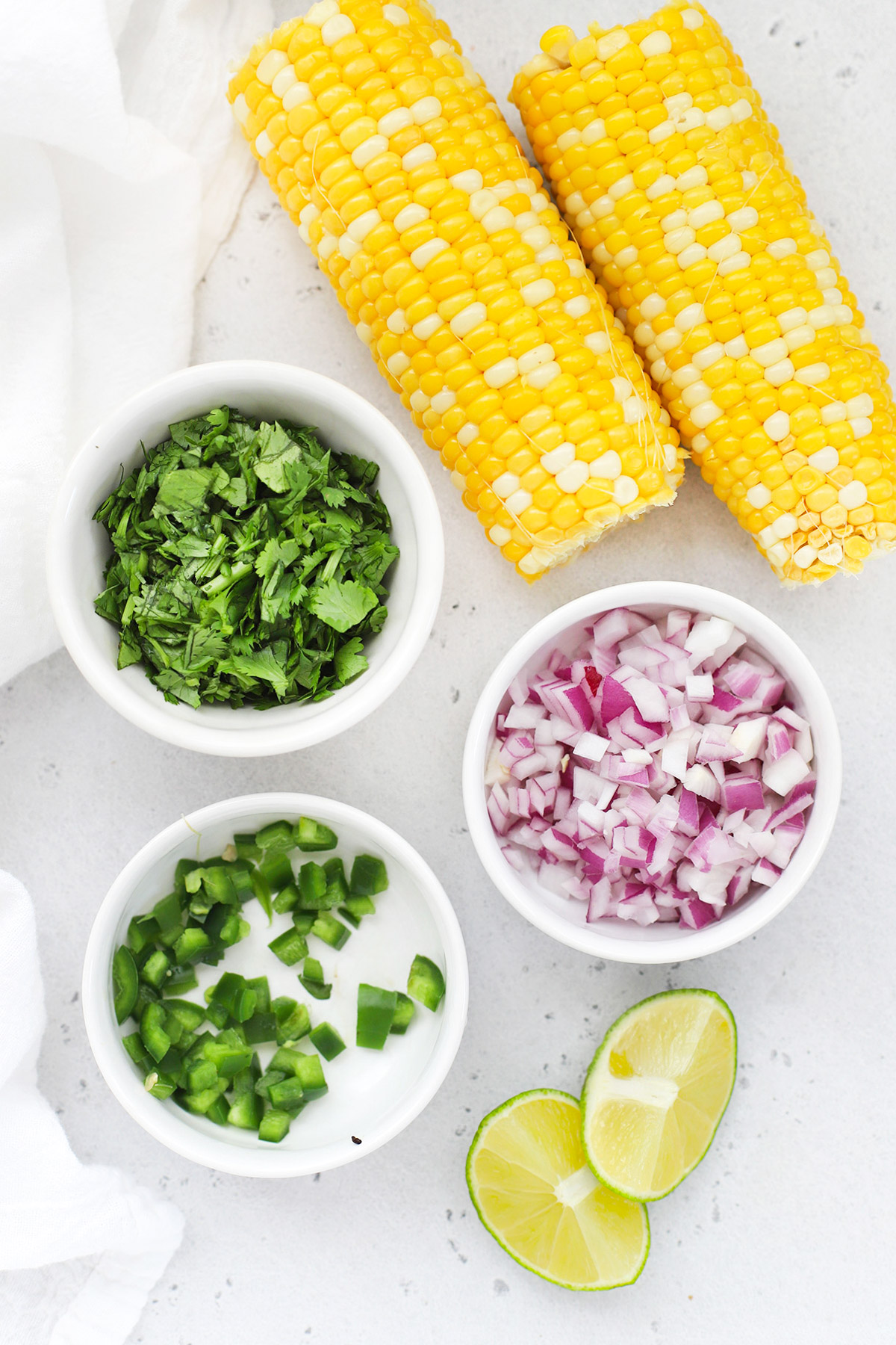 Overhead view of ingredients for fresh corn salsa arranged on a white backdrop--corn on the cob, cilantro, red onion, jalapeno, and fresh lime