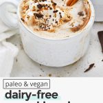 Front view of a steaming mug of dairy-free pumpkin spice hot chocolate topped with coconut whipped cream, shaved chocolate, and pumpkin spice with text overlay that reads "paleo & vegan pumpkin spice hot chocolate: cozy + creamy + amazing!"