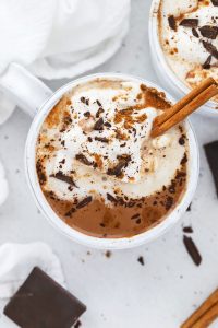 Overhead view of a steaming mug of dairy-free pumpkin spice hot chocolate topped with coconut whipped cream, shaved chocolate, and pumpkin spice