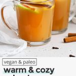 Front view of glass mugs of hot spiced cider garnished with apple slices, orange slices, and cinnamon sticks with text overlay that reads "vegan & paleo warm & cozy spiced apple cider: easy + soothing + festive"