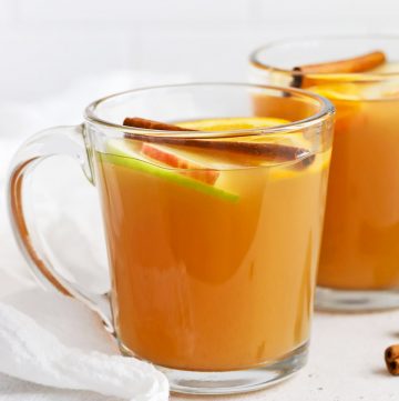 Front view of glass mugs of hot spiced cider garnished with apple slices, orange slices, and cinnamon sticks